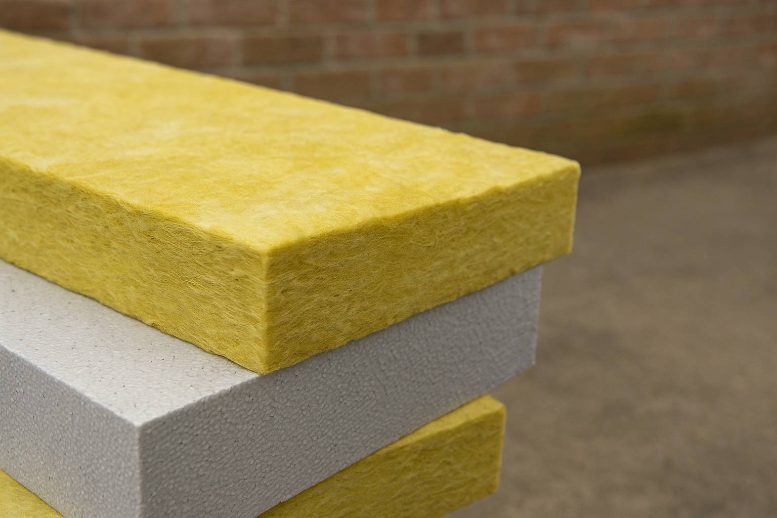 Trzy warstwy materiałów izolacyjnych ułożone jedna na drugiej — na górze żółta wełna mineralna, poniżej biały styropian, a na samym dole kolejna warstwa żółtej wełny; tło stanowi rozmyta ceglana ściana i betonowa posadzka.